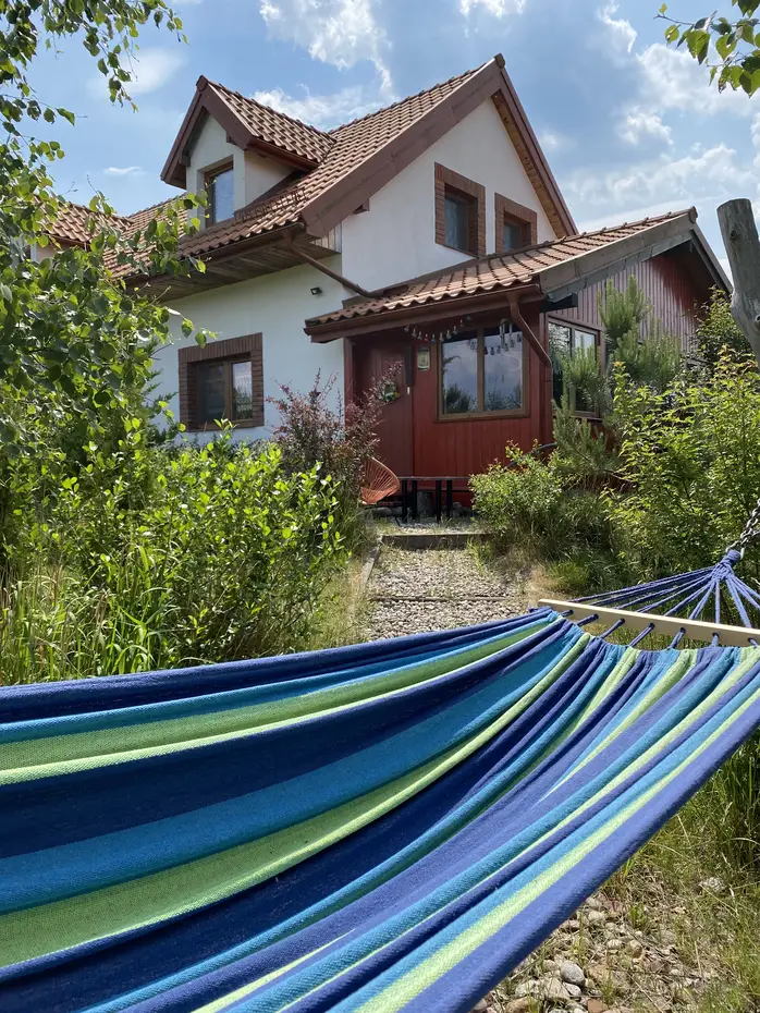 Kolorowy hamak w odcieniach niebieskiego i zielonego rozwieszony w ogrodzie przed jednorodzinnym domem z czerwonym dachem i drewnianą werandą, otoczonym bujną zielenią w słoneczny dzień