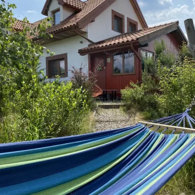 IMG_0596 Kolorowy hamak w odcieniach niebieskiego i zielonego rozwieszony w ogrodzie przed jednorodzinnym domem z czerwonym dachem i drewnianą werandą, otoczonym bujną zielenią w słoneczny dzień